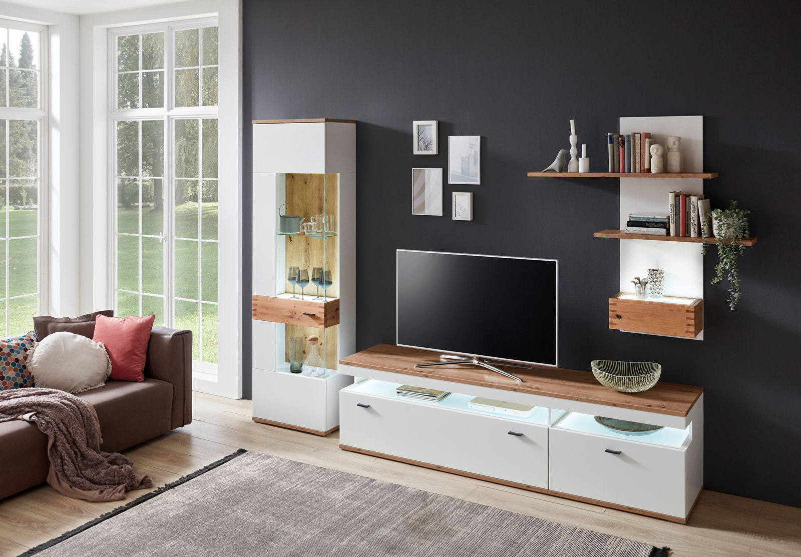 Moderne Wandkombination in einem Wohnzimmer mit TV, Vitrine und Regalen, aus hellem Holz und weißen Elementen, seitliche Perspektive.