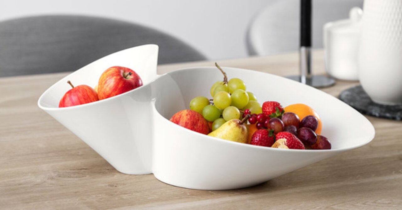 Weiße, doppelte Schale in moderner Form, gefüllt mit frischem Obst, auf einem Holztisch aus seitlicher Perspektive.
