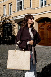 Beiger Shopper mit dezentem Muster und zwei Henkeln, getragen von einer Frau im Freien; Perspektive: leicht seitliche Ansicht auf die Tasche.