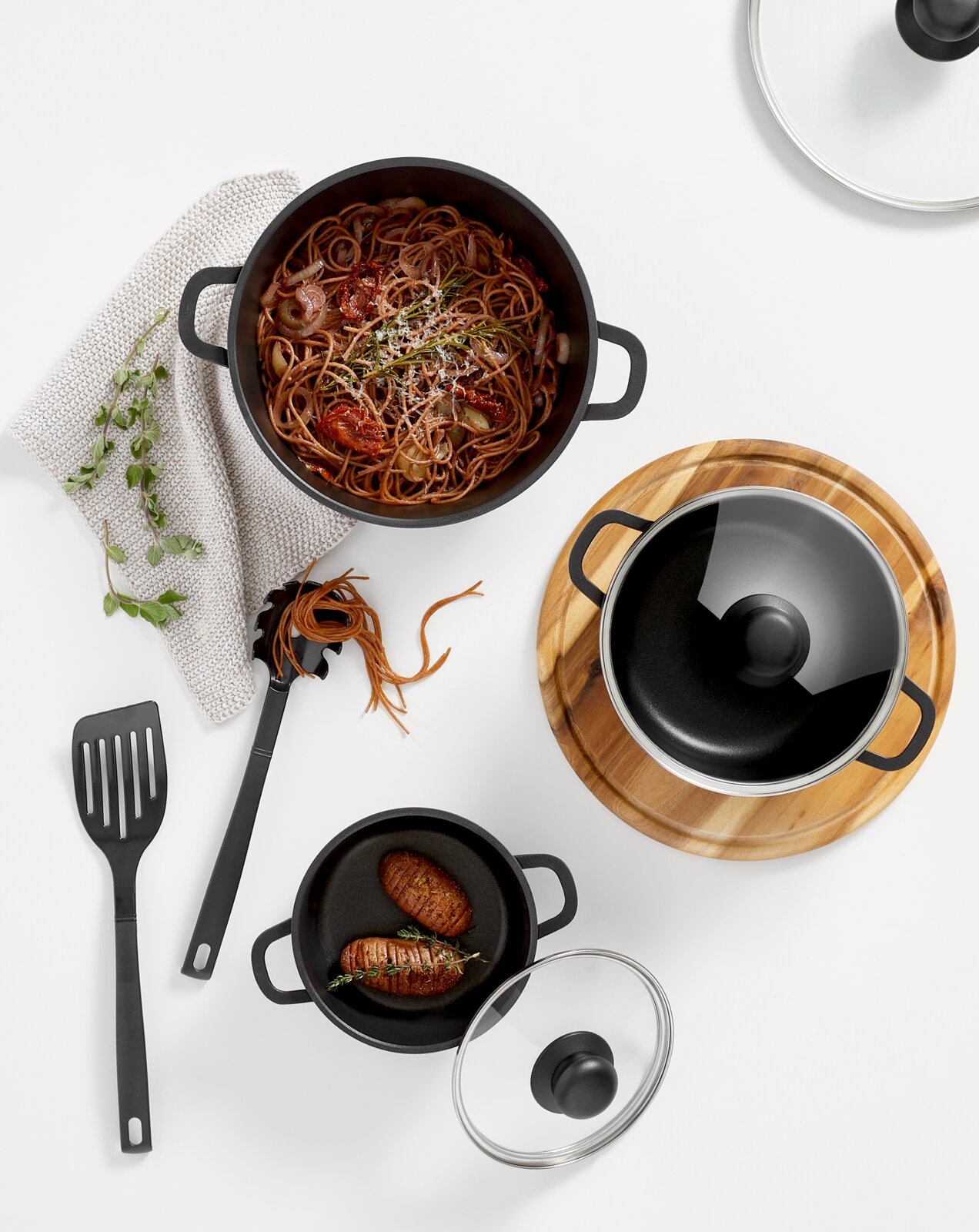 Bovenaanzicht van een 3-delige pannenset met een grote pan vol spaghetti, een kleinere pan met twee gebakken aardappelen en nog een pan met glazen deksel op een houten onderzetter. Ook zijn er keukengerei en kruiden te zien.