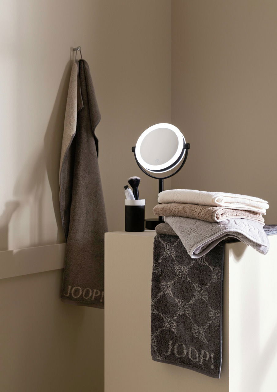 Standkosmetikspiegel mit Touch-Funktion, beleuchtet, auf einem Tisch neben Handtüchern und einem Becher mit Kosmetikpinseln, seitliche Perspektive