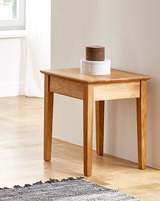 Schlichter Beistelltisch aus hellem Holz mit quadratischer Tischplatte in einer hellen Zimmerecke, fotografiert aus leicht erhöhter schräger Vorderansicht.