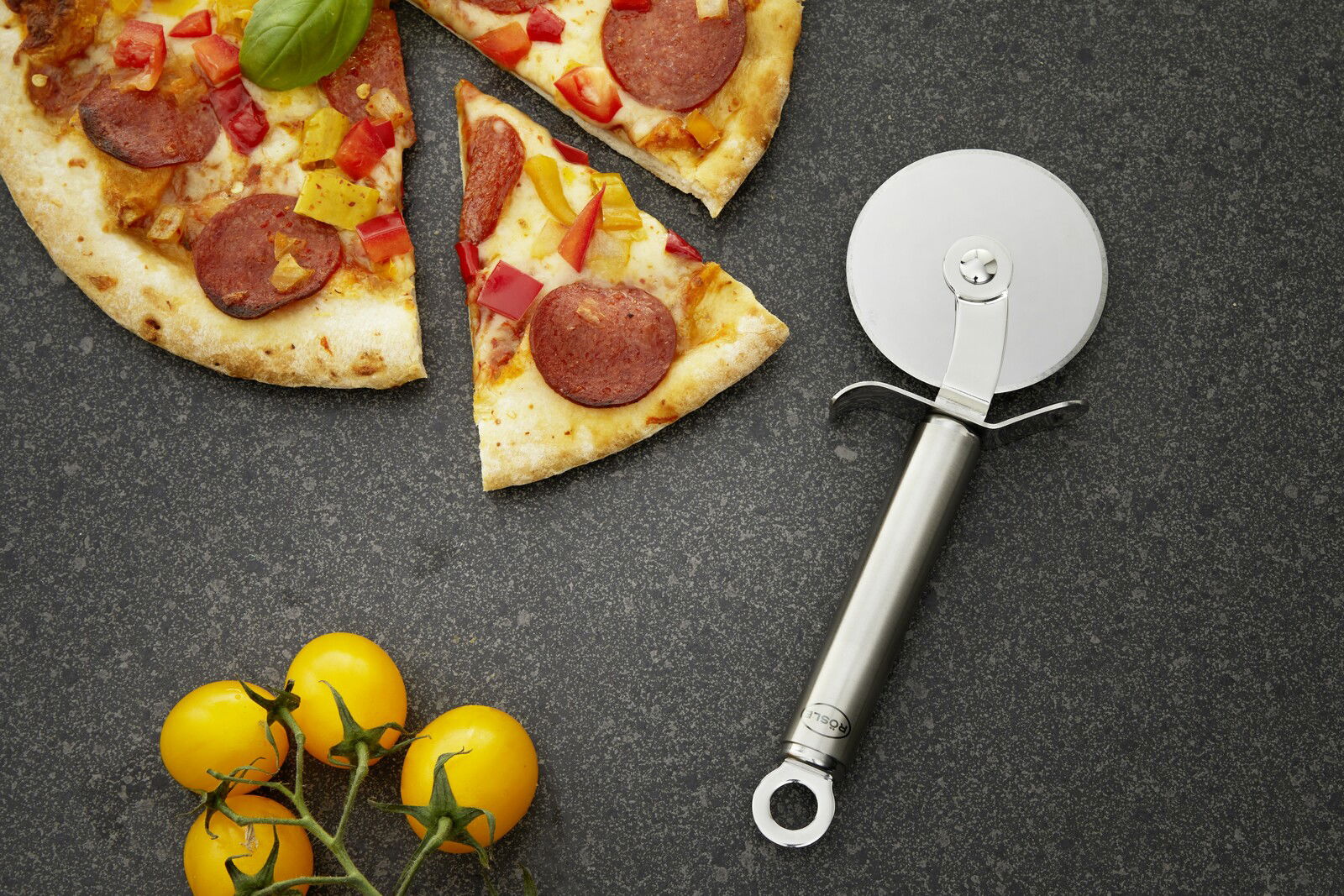 Pizzaschneider aus Edelstahl neben einer angeschnittenen Pizza mit Peperoni und Paprika, von oben betrachtet.