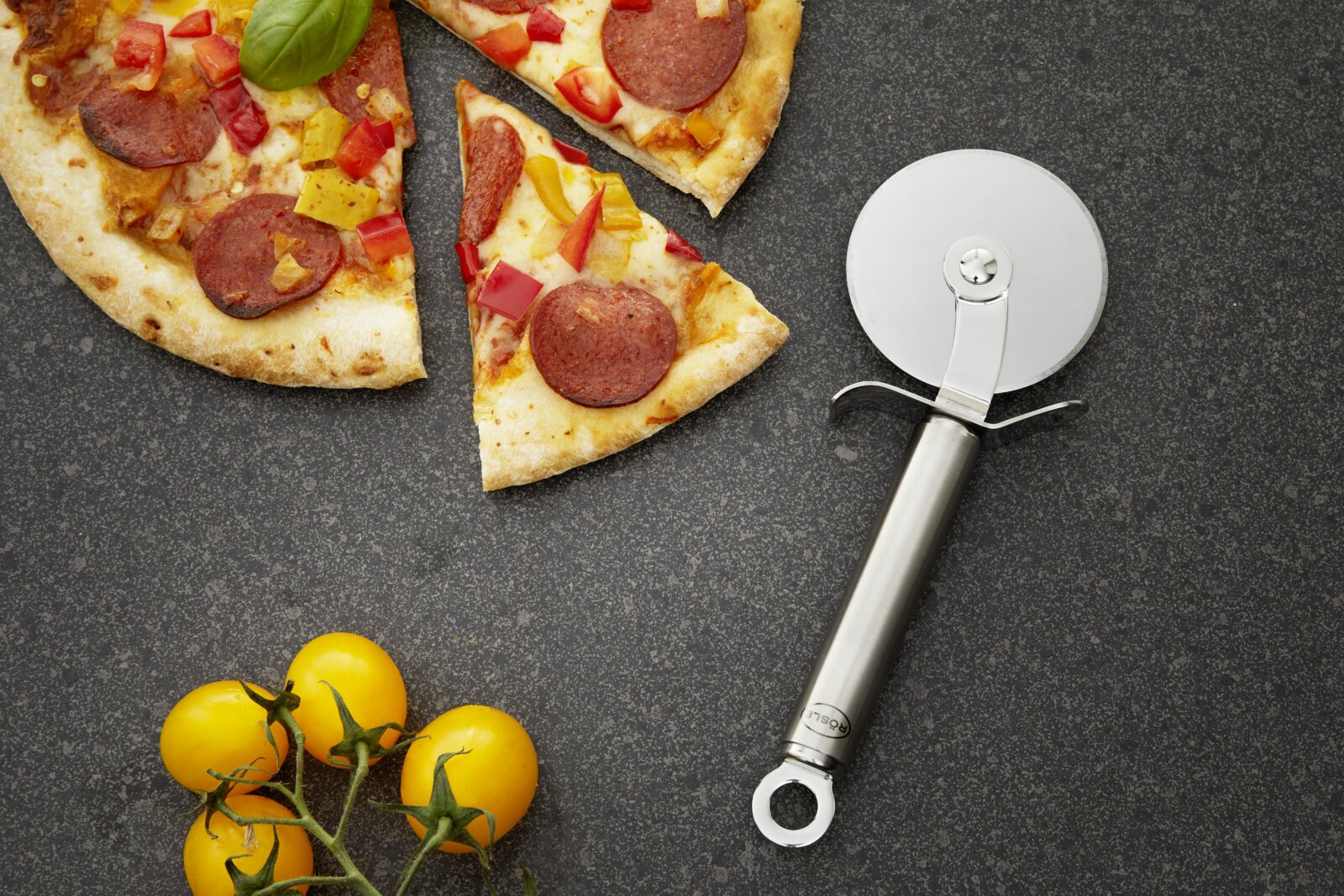 Pizzaschneider aus Edelstahl neben einer angeschnittenen Pizza mit Peperoni und Paprika, von oben betrachtet.