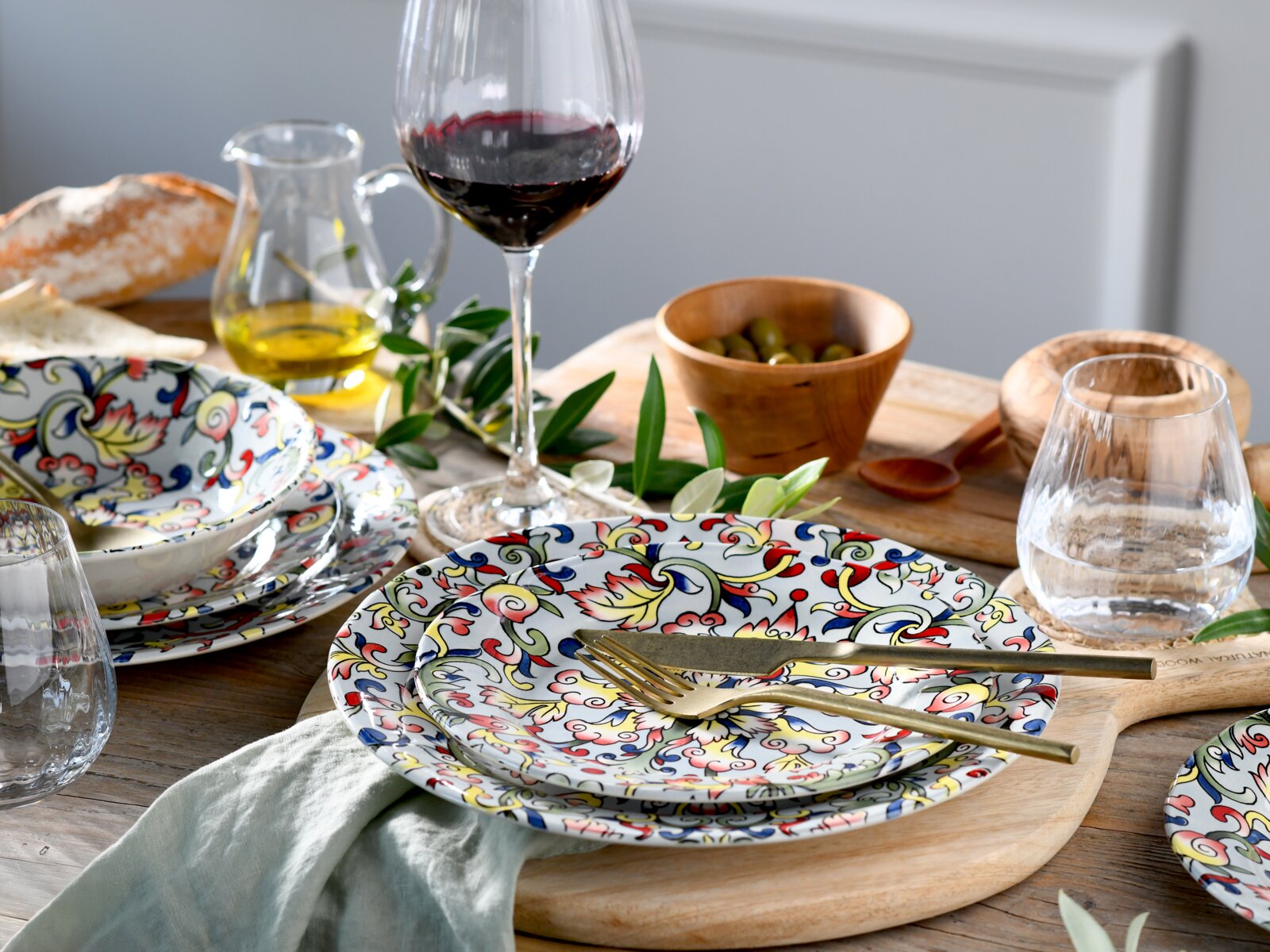 Schräg von oben fotografiertes, mediterranes Tisch-Arrangement mit buntem Keramikgeschirr im floralen Muster, goldfarbenem Besteck, Rotweinglas, Wasserglas, Oliven in Holzschale, Olivenölkanne und Brot auf Holztisch.