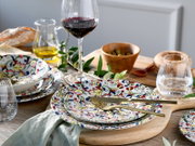Schräg von oben fotografiertes, mediterranes Tisch-Arrangement mit buntem Keramikgeschirr im floralen Muster, goldfarbenem Besteck, Rotweinglas, Wasserglas, Oliven in Holzschale, Olivenölkanne und Brot auf Holztisch.