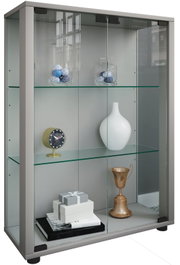 Graue Glasvitrine mit zwei Glasböden und Schiebetüren, befüllt mit Deko (Vasen, Uhr, Pokal, Geschenkboxen), in leicht schräger Frontansicht von links.