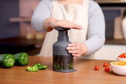 Grijze handmatige groentesnijder/food chopper met drukmechanisme, vastgehouden door een persoon op een houten aanrecht met paprika en cherrytomaten; frontaal aanzicht op werkhoogte
