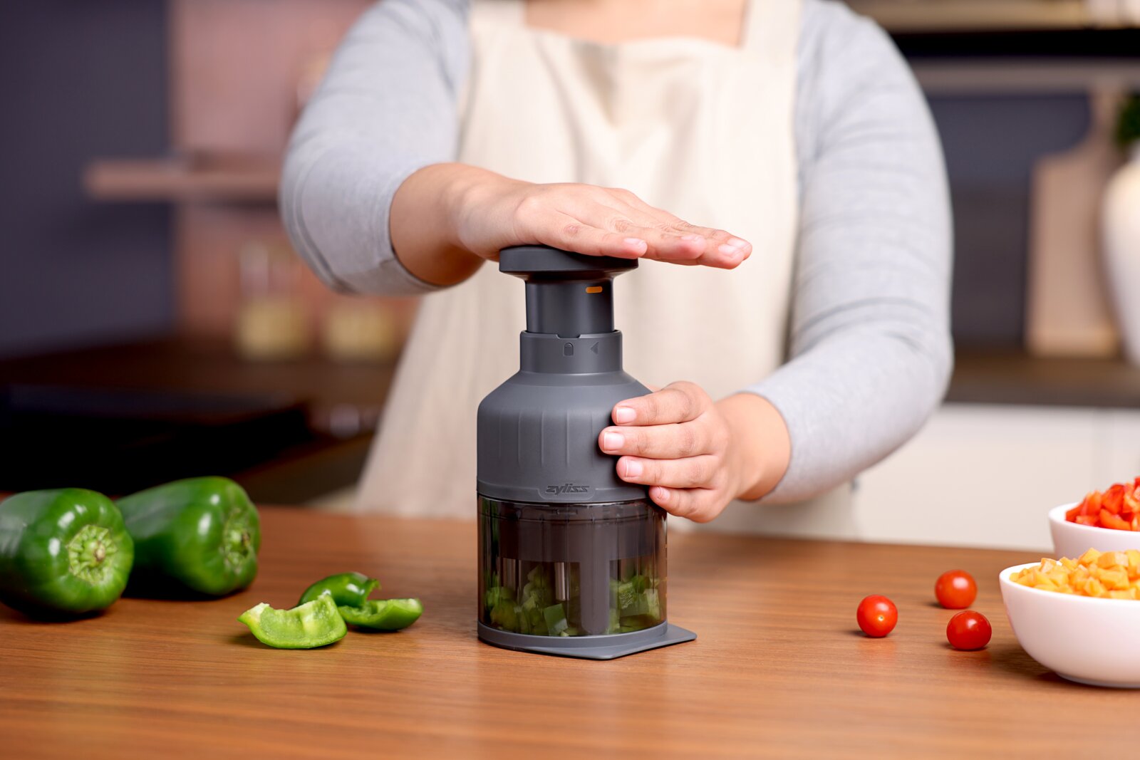Grijze handmatige groentesnijder/food chopper met drukmechanisme, vastgehouden door een persoon op een houten aanrecht met paprika en cherrytomaten; frontaal aanzicht op werkhoogte