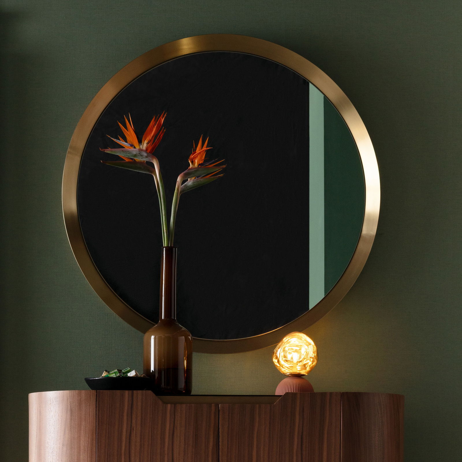 Runder Wandspiegel mit goldfarbenem Rahmen, reflektiert eine Vase mit exotischen Blumen, auf einem Holzschrank stehend. Frontalansicht.