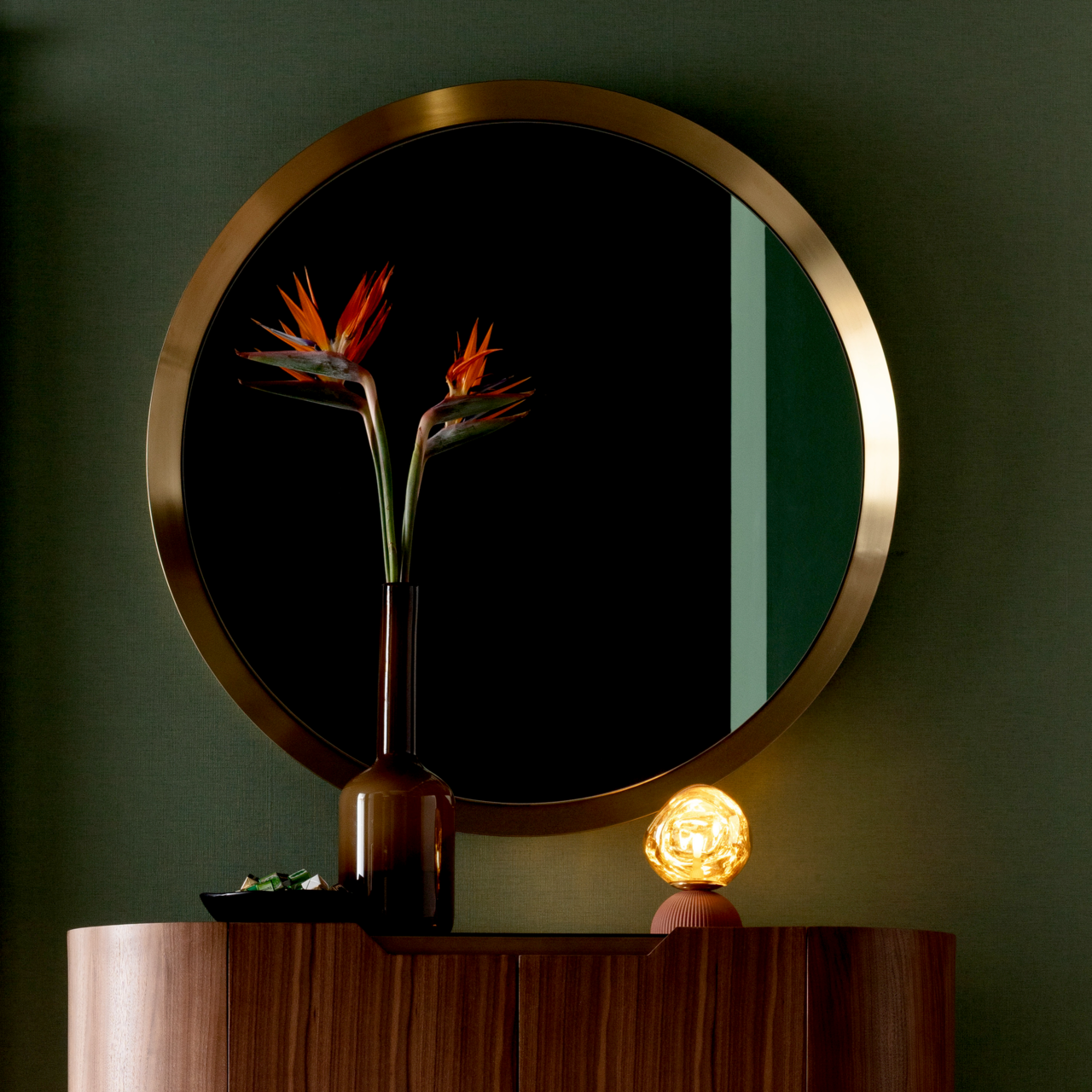 Runder Wandspiegel mit goldfarbenem Rahmen, reflektiert eine Vase mit exotischen Blumen, auf einem Holzschrank stehend. Frontalansicht.