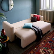 Beiges Funktionssofa PONTE in einem gemütlichen Wohnzimmer, ausgeklappt als Bett mit bunten Kissen und Decke, seitliche Perspektive.