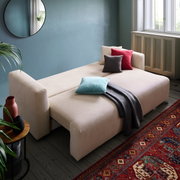 Beiges Funktionssofa PONTE in einem gemütlichen Wohnzimmer, ausgeklappt als Bett mit bunten Kissen und Decke, seitliche Perspektive.