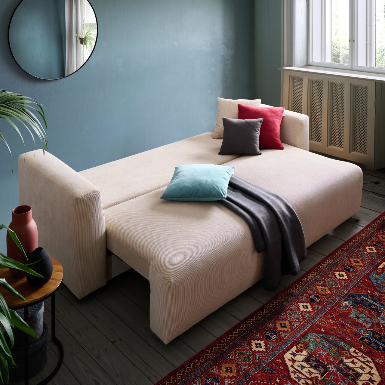 Beiges Funktionssofa PONTE in einem gemütlichen Wohnzimmer, ausgeklappt als Bett mit bunten Kissen und Decke, seitliche Perspektive.