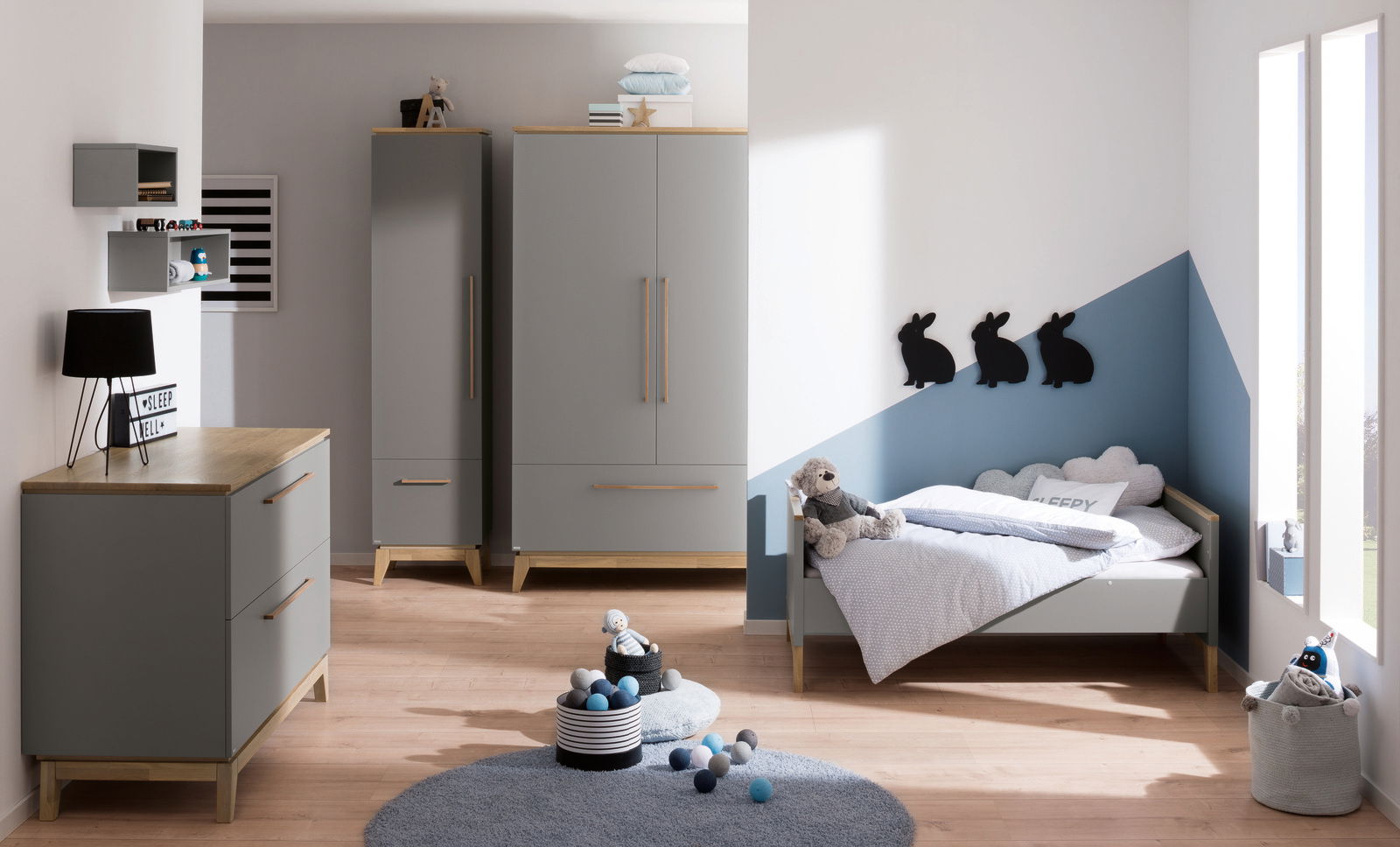 Kinderzimmer mit grauem Kleiderschrank, Kommode und Bett, dekoriert mit Kuscheltieren und Wanddekorationen in Hasenform. Perspektive von vorne.