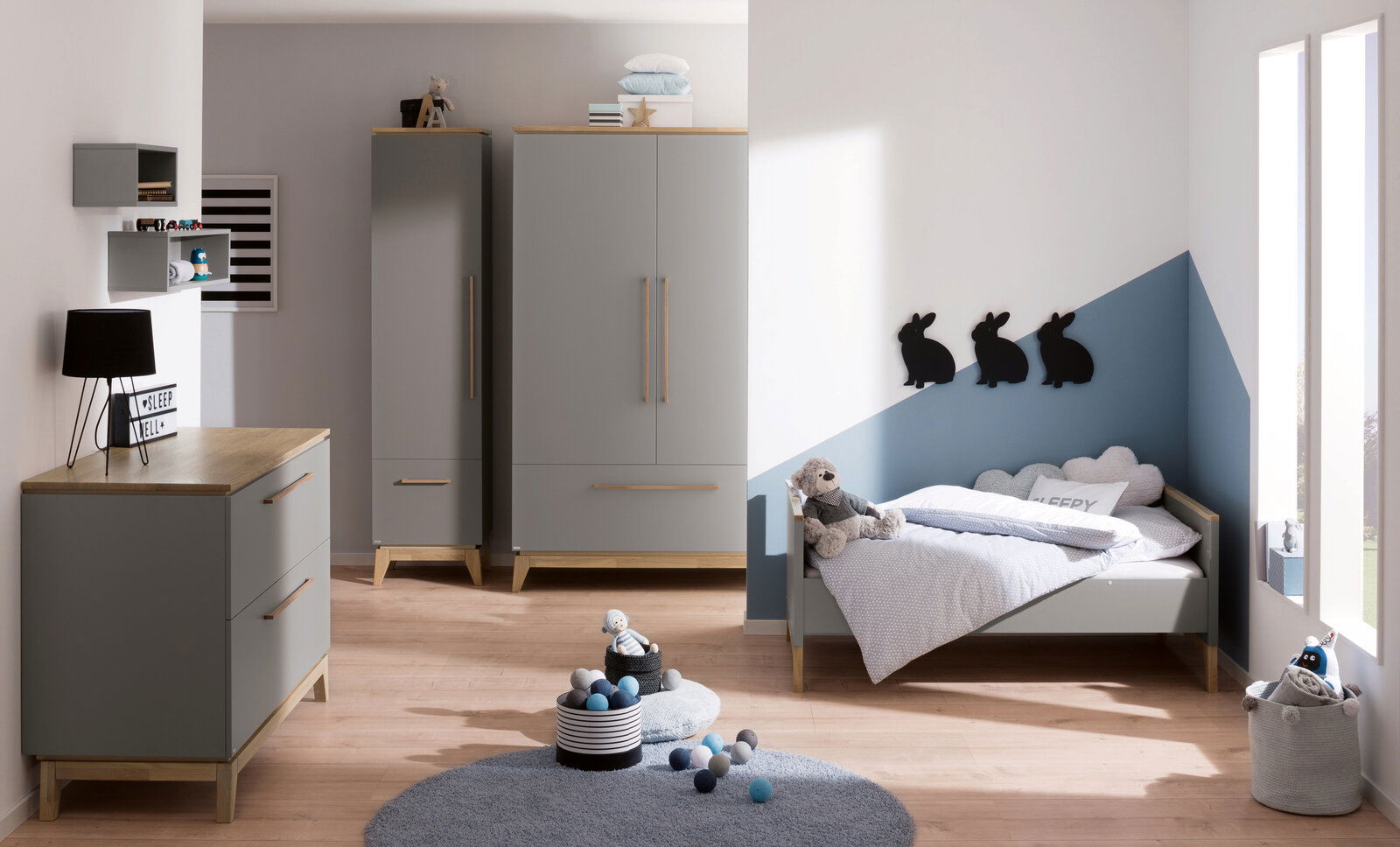 Kinderzimmer mit grauem Kleiderschrank, Kommode und Bett, dekoriert mit Kuscheltieren und Wanddekorationen in Hasenform. Perspektive von vorne.