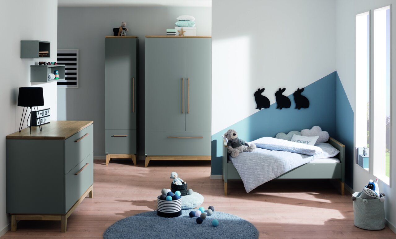 Kinderzimmer mit grauem Kleiderschrank, Kommode und Bett, dekoriert mit Kuscheltieren und Wanddekorationen in Hasenform. Perspektive von vorne.
