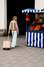 Beiger Einkaufstrolley mit ausziehbarem Griff und dezentem Muster, von einer Frau auf dem Gehweg neben einem blau-weiß gestreiften Obststand gezogen, Seitenansicht in leichter Schrägperspektive.