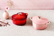 Zwei herzförmige Le Creuset Gusseisen-Bräter mit Deckel, einer in Rot und einer in zartem Rosa, auf heller Küchenarbeitsplatte vor rosafarbener Fliesenwand, leicht schräg von vorn fotografiert.