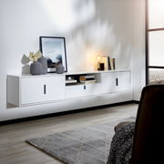 Modernes weißes TV-Hängeboard mit drei Türen und einem offenen Fach, dekoriert mit Vasen, Büchern und einer Lampe, seitliche Perspektive.