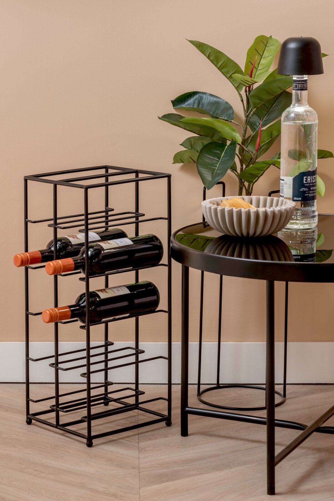 Schwarzes Weinregal aus Metall mit Platz für mehrere Flaschen, seitliche Perspektive, neben einem Tisch mit Pflanze und Getränk.