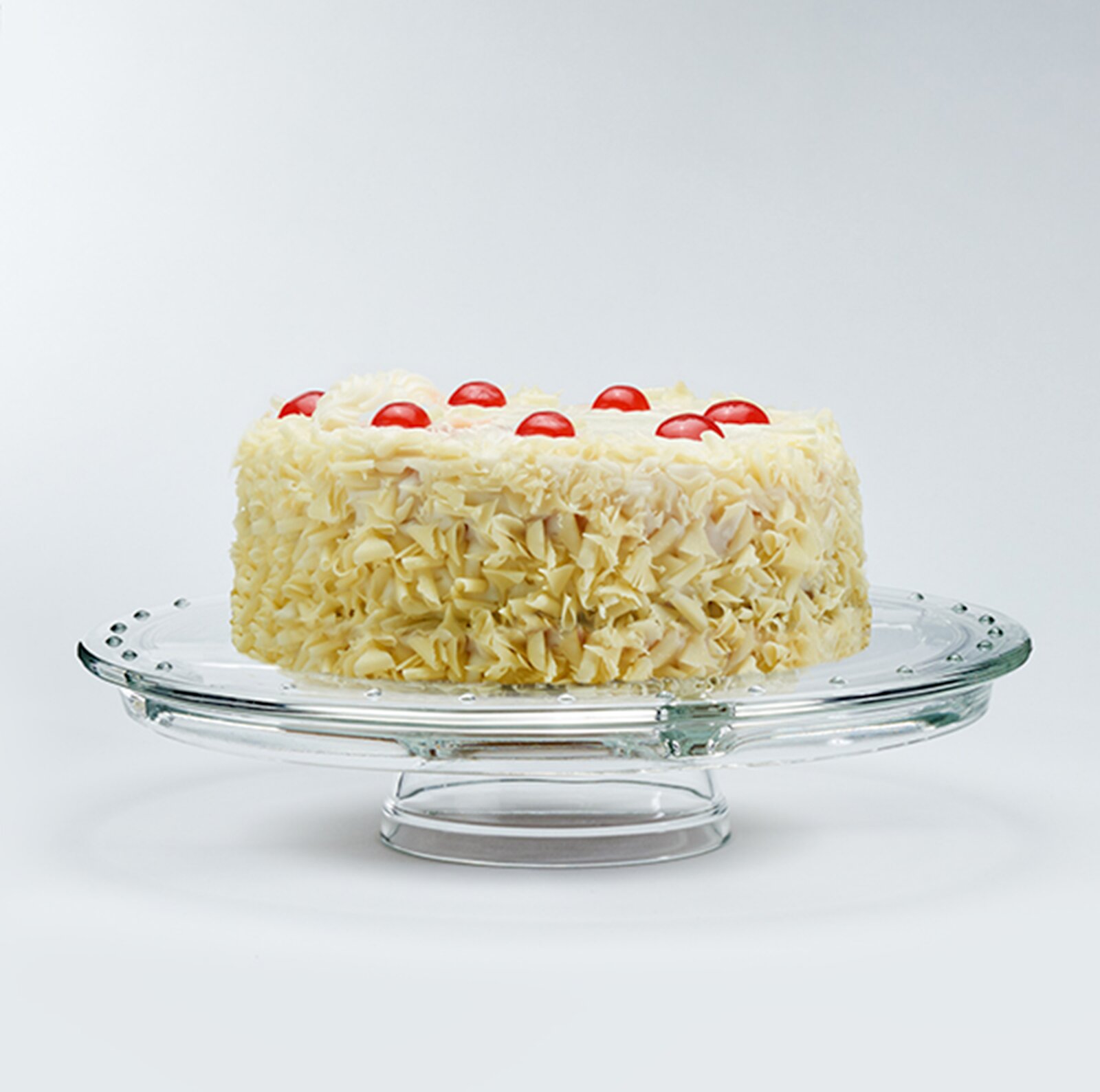 Runde Torte mit weißer Creme, rundum mit weißen Schokoraspeln bedeckt und mit roten Kirschen dekoriert, auf einer Glas-Tortenplatte vor hellem Hintergrund, Seitenansicht leicht von oben