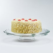 Runde Torte mit weißer Creme, rundum mit weißen Schokoraspeln bedeckt und mit roten Kirschen dekoriert, auf einer Glas-Tortenplatte vor hellem Hintergrund, Seitenansicht leicht von oben
