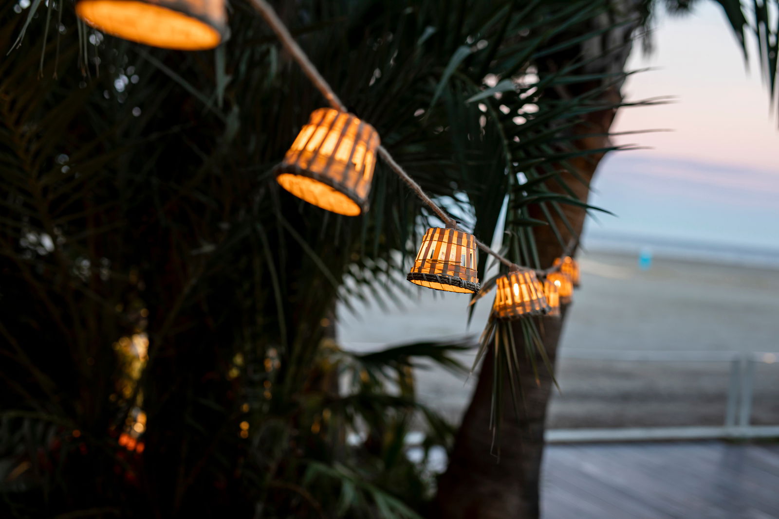 Lichterkette mit kleinen, leuchtenden Lampenschirmen aus Holz, aufgehängt zwischen Palmen mit Blick auf einen Strand bei Sonnenuntergang.