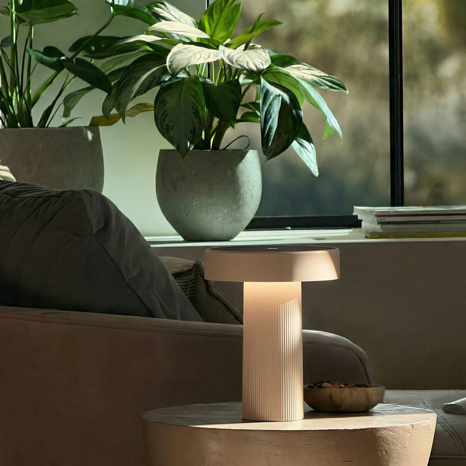 Moderne, zylindrische Tischlampe in Beige mit warmem Licht auf einem runden Couchtisch neben einem Sofa, im Hintergrund Zimmerpflanzen am Fenster – Perspektive: frontale leicht seitliche Ansicht auf Lampenschirm und Sockel
