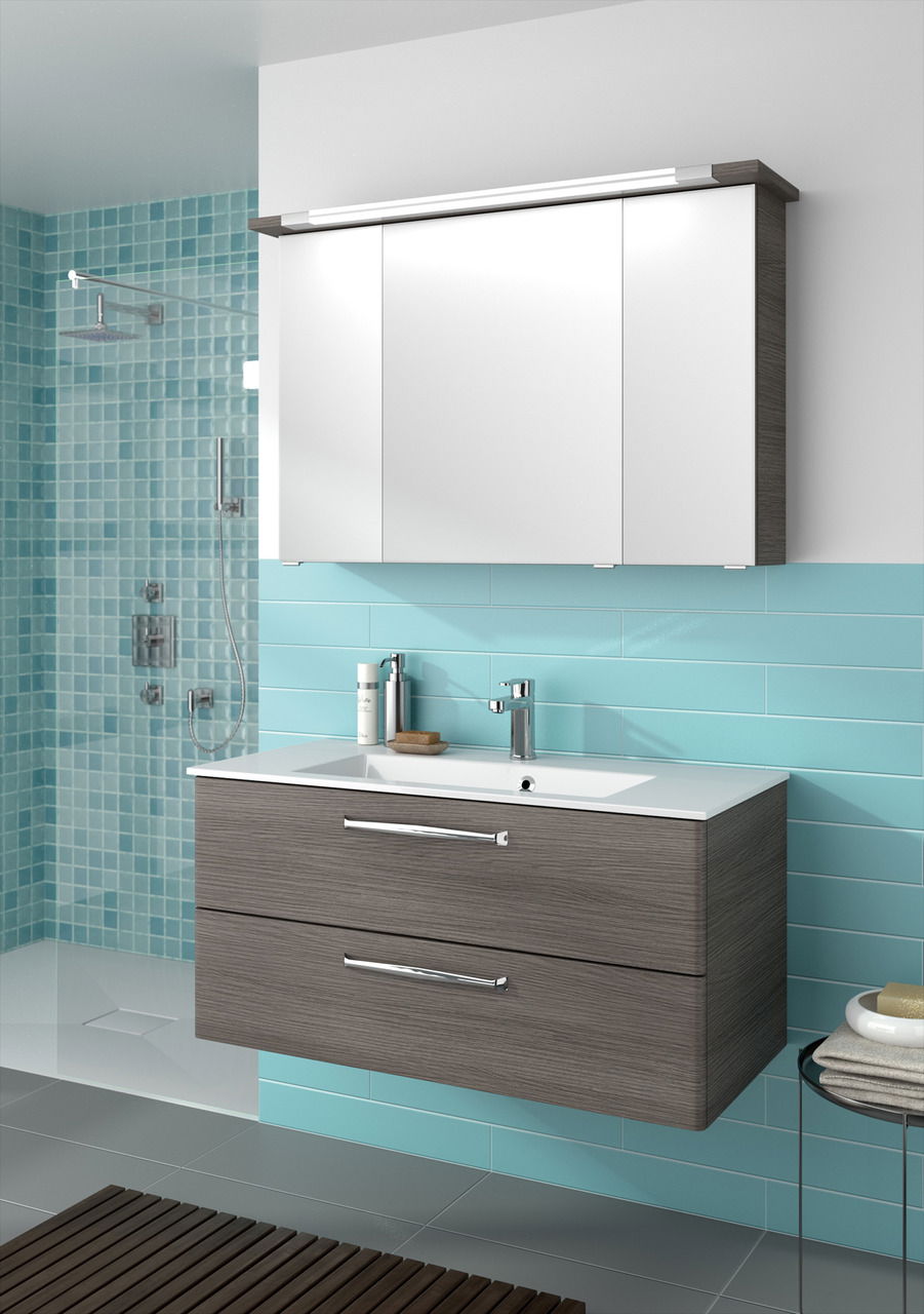 Moderner Badblock mit weißem Waschbecken und zwei Schubladen in Holzoptik, vor türkisfarbenen Fliesen, aus seitlicher Perspektive.