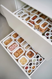 Een bovenaanzicht van een geopend ladesysteem met witte, rechthoekige voorraadbakken, gevuld met diverse levensmiddelen zoals pasta, koekjes en ontbijtgranen.