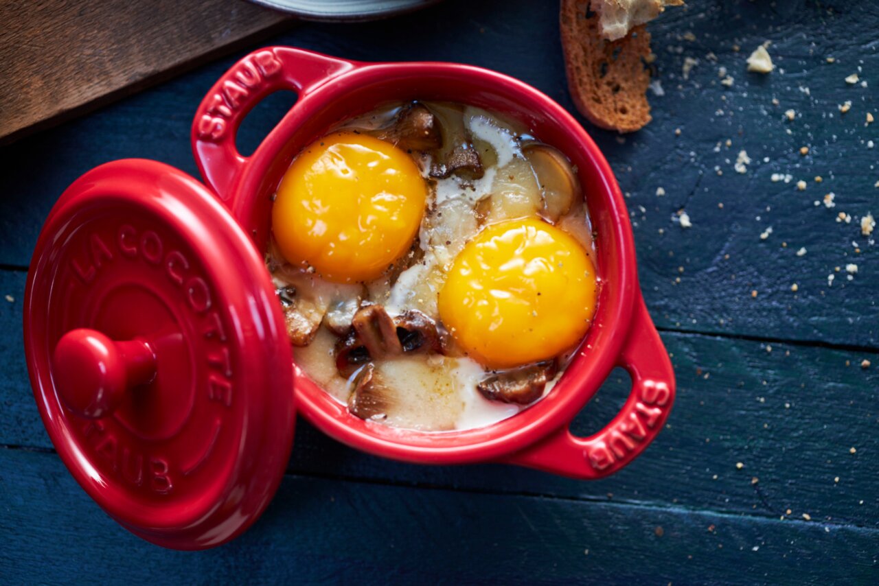 Rote Mini-Cocotte mit geöffnetem Deckel, gefüllt mit zwei Eigelb und Pilzen, aus der Vogelperspektive fotografiert.