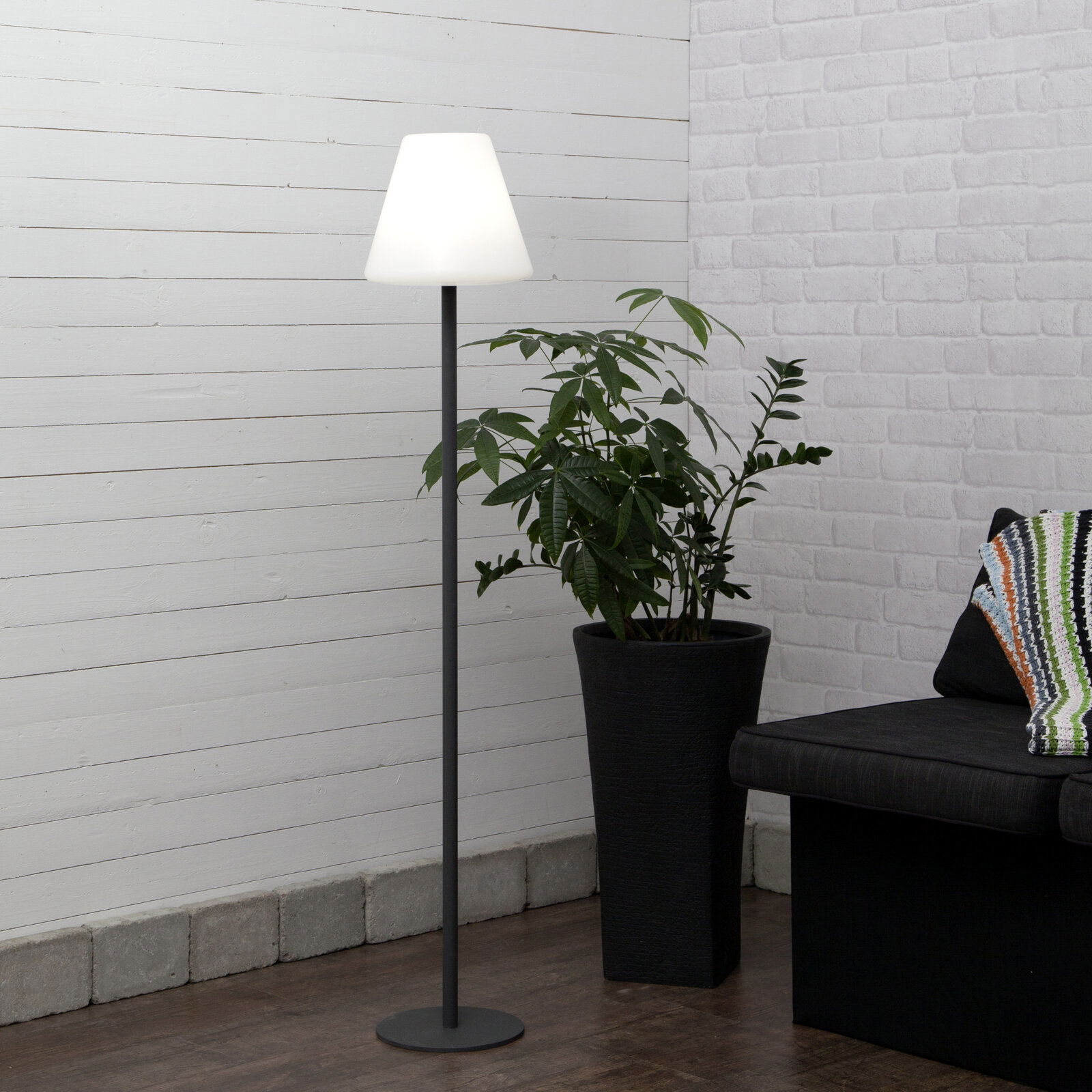 STAR TRADING KRETA buitenstaande lamp Moderne Stehlampe mit schwarzem Metallfuß und weißem, konischem Lampenschirm in einer Wohnzimmerecke neben großer Zimmerpflanze und Sofa, frontale Perspektive
