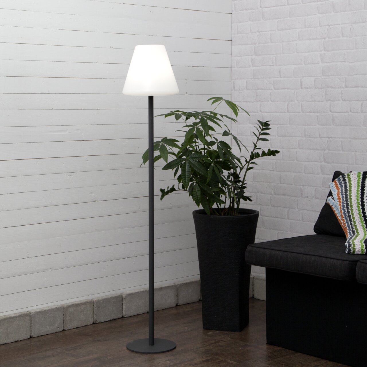 STAR TRADING Außen-Stehleuchte KRETA Moderne Stehlampe mit schwarzem Metallfuß und weißem, konischem Lampenschirm in einer Wohnzimmerecke neben großer Zimmerpflanze und Sofa, frontale Perspektive