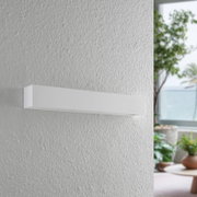 Moderne weiße Wandleuchte in rechteckiger Form, seitliche Perspektive, montiert an einer strukturierten Wand in einem hellen Raum mit Pflanzen im Hintergrund.