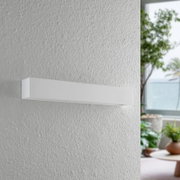 Moderne weiße Wandleuchte in rechteckiger Form, seitliche Perspektive, montiert an einer strukturierten Wand in einem hellen Raum mit Pflanzen im Hintergrund.