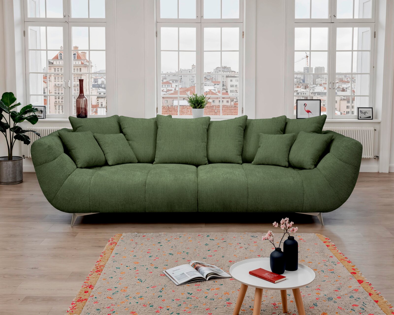 Großes grünes Stoffsofa mit vielen Kissen und Metallfüßen im hellen Wohnzimmer vor großen Fenstern, Frontalansicht auf Augenhöhe.