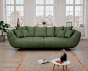 Großes grünes Stoffsofa mit vielen Kissen und Metallfüßen im hellen Wohnzimmer vor großen Fenstern, Frontalansicht auf Augenhöhe.