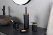 Modernes Bad-Accessoire-Set in Schwarz mit geriffelter Oberfläche und Bambusdetails (Toilettenbürstenhalter, Seifenspender, Zahnputzbecher und Seifenschale) auf dunkler Ablage neben Steinwaschbecken, Frontansicht in leichter Seitenperspektive.
