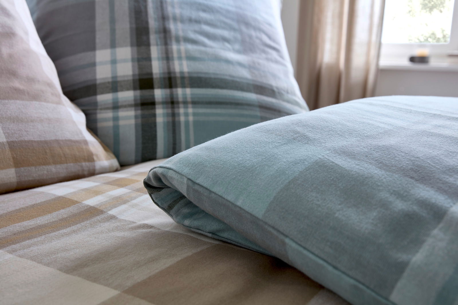 Nahaufnahme von Bettwäsche mit kariertem Muster in Blau- und Beigetönen, auf einem Bett arrangiert.