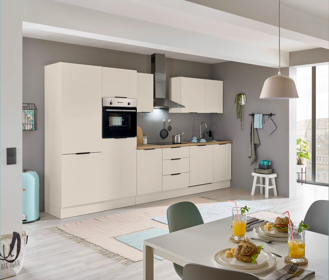 Moderne Einbauküche in heller Farbe mit integriertem Backofen und Kühlschrank, Dunstabzugshaube und Arbeitsplatte aus Holz. Perspektive von der Seite mit Blick auf den Esstisch im Vordergrund.