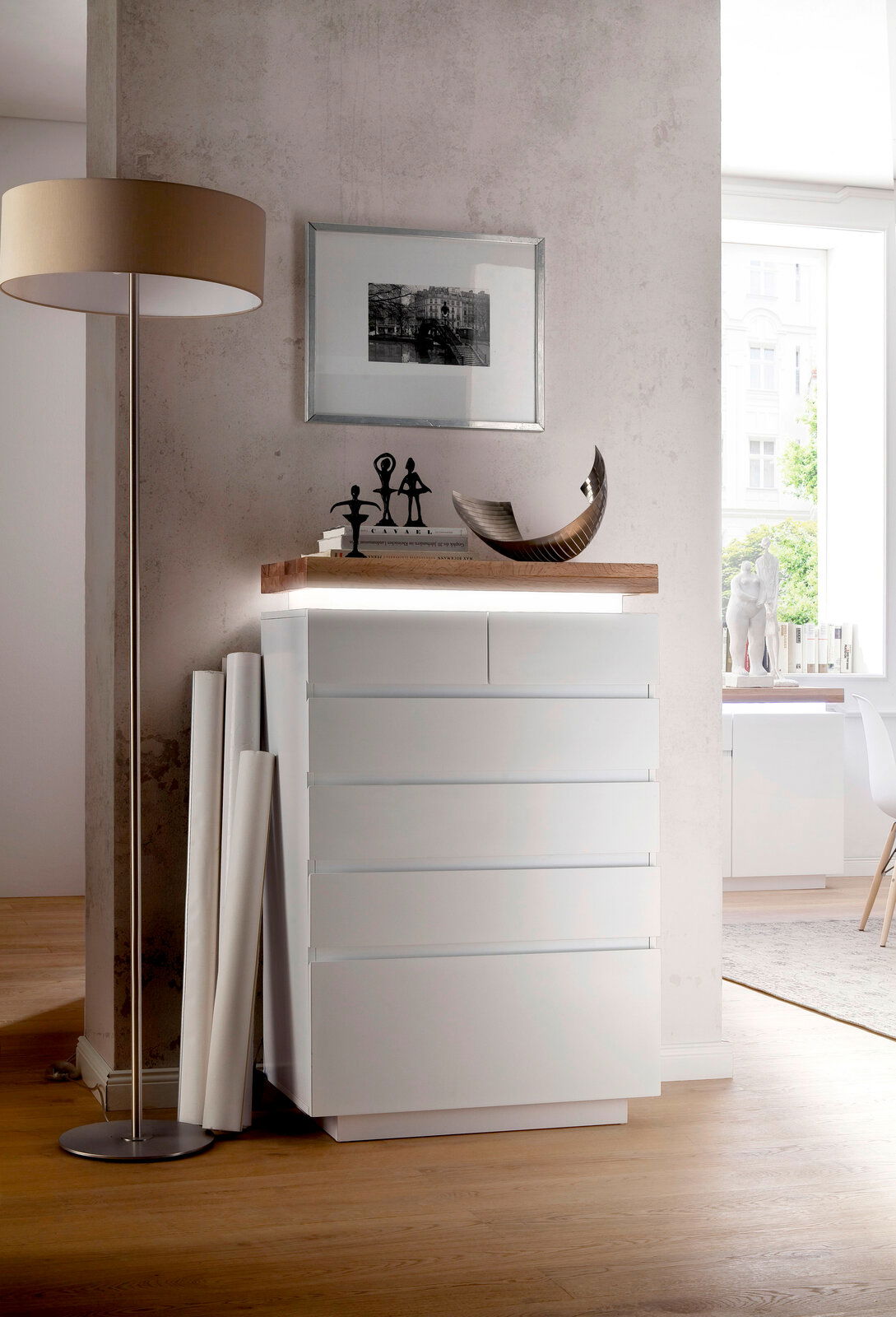 Novel Hochkommode ROMINA Moderne weiße Hochkommode mit fünf Schubladen, seitliche Perspektive, dekoriert mit Skulpturen und Büchern auf der Oberseite.