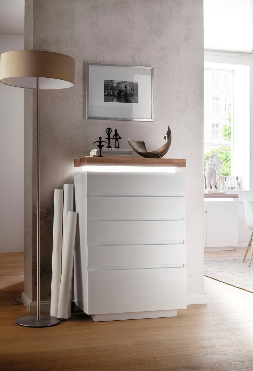 Moderne weiße Hochkommode mit fünf Schubladen, seitliche Perspektive, dekoriert mit Skulpturen und Büchern auf der Oberseite.