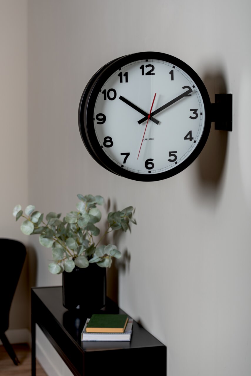 Wanduhr im klassischen Design mit schwarzem Rahmen und weißen Zifferblatt, seitliche Perspektive, montiert an einer hellen Wand über einem Tisch mit Pflanzen und Büchern.