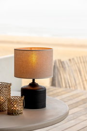 Tischlampe mit beigem Stoffschirm und schwarzem, geripptem Sockel auf einem Tisch, seitliche Perspektive.