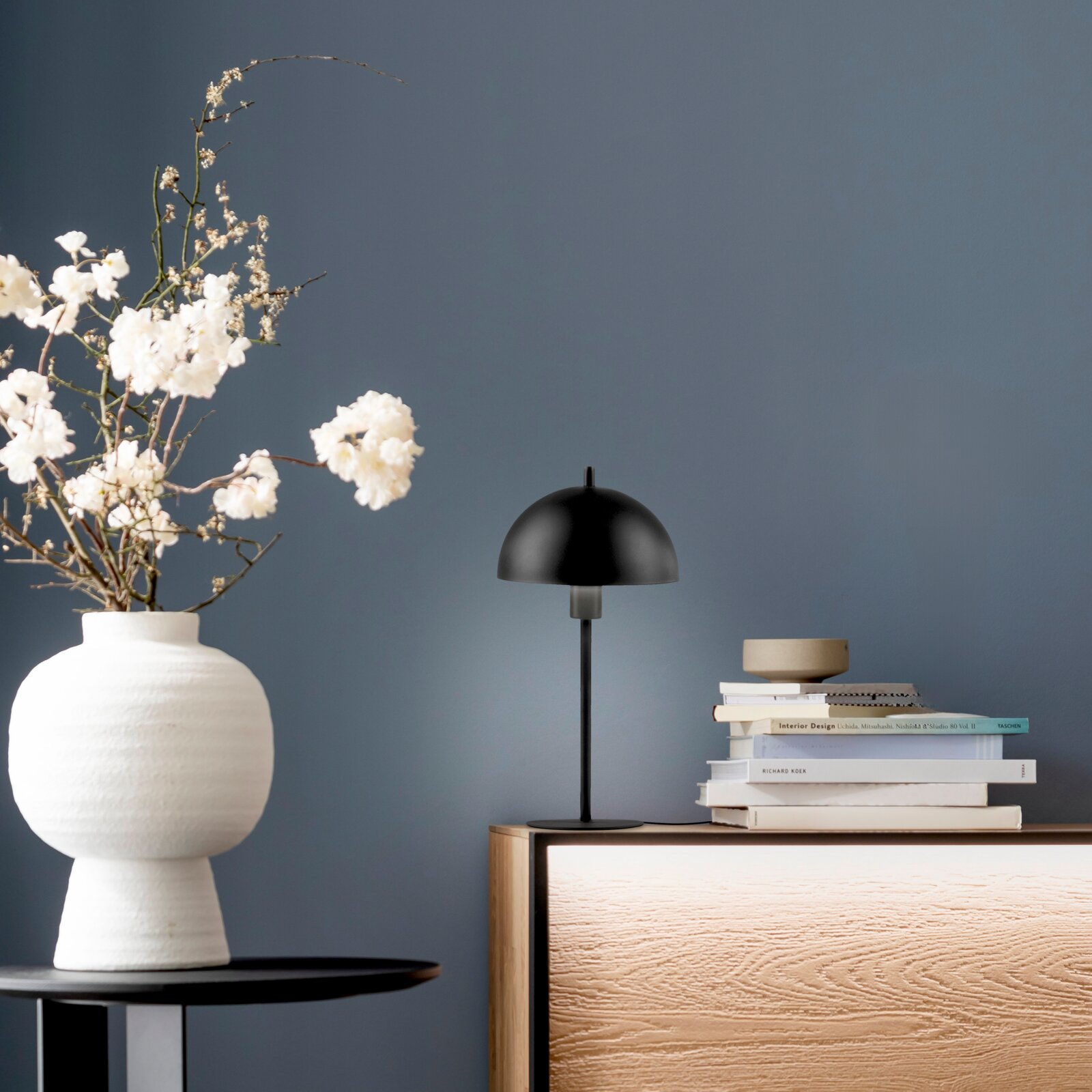 Schwarze Tischlampe mit halbrundem Schirm auf einem Holzsideboard neben einem Bücherstapel, links eine weiße Vase mit weißen Blüten vor graublauer Wand, Frontansicht auf Augenhöhe.