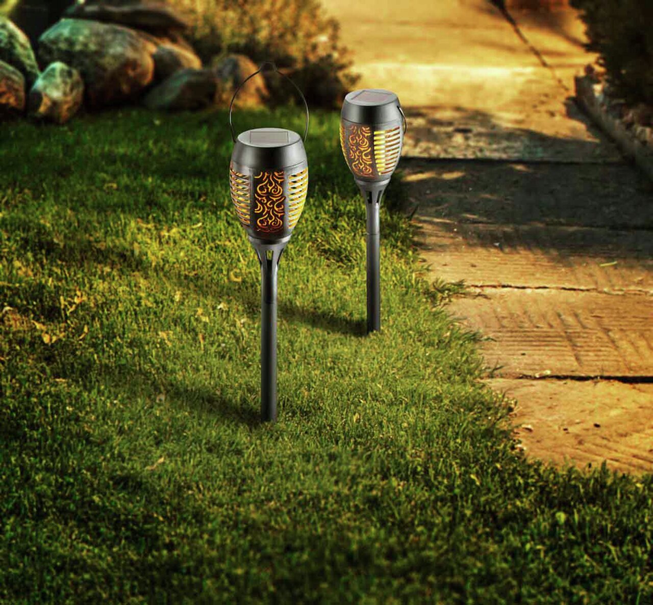 Ambia Garden Solarleuchte Flame II Zwei Solarleuchten Flame II mit flammenähnlichem Lichtmuster, auf einem Rasen neben einem Gehweg platziert, aus seitlicher Perspektive fotografiert.