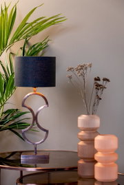 Moderner Lampenschirm in Zylinderform mit schwarzem Stoff, auf einem eleganten Metallfuß, umgeben von Pflanzen und dekorativen Vasen, seitliche Perspektive.