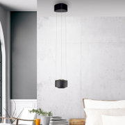 Moderne schwarze Pendelleuchte mit zylindrischer Deckenhalterung und kompaktem Lampenschirm an drei dünnen Seilen, frontal in leichter Untersicht vor heller Betonwand im Wohnraum