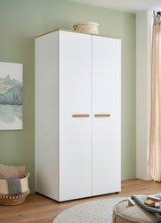 Weißer zweitüriger Kleiderschrank mit Holzoptik-Oberplatte und Holzgriffen, im Zimmer vor grüner Wand – frontale Ansicht mit leichter Perspektive von links.